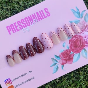 May include: A collection of press-on nails in different styles. Some nails are a deep burgundy with white polka dots, others are light pink with burgundy polka dots, and some are a nude colour. The nails are displayed on a pink card with the text "PRESSONNAILS by del".