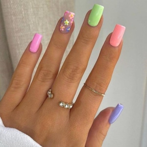 May include: A hand displaying long, square-shaped nails in pastel shades of pink, green, peach, and lilac. One nail is decorated with tiny floral designs on a clear base. Several delicate rings adorn the fingers.