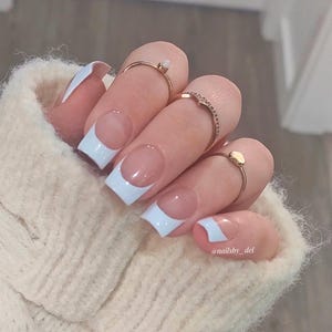 May include: Close-up of a hand with white square-shaped nails with a white french tip. The hand is wearing several gold rings. The hand is resting on a cream-coloured fuzzy surface. The text "@nailsby_del" is visible in the bottom right corner of the image.