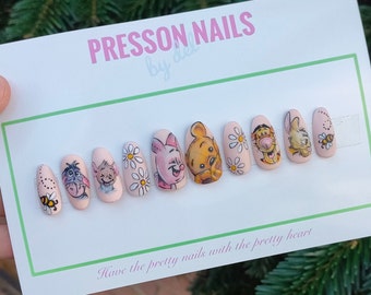 May include: A set of press-on nails featuring cartoon characters from Winnie the Pooh. The nails are painted light pink with illustrations of Eeyore, Piglet, Winnie the Pooh, Tigger, and other designs. The text "PRESSON NAILS" is visible.