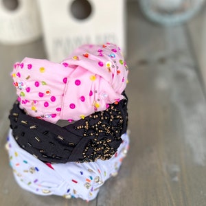 May include: Three knotted headbands with different embellishments. The top headband is pink with pink and multi-colored beads. The middle headband is black with gold beads in a spiderweb design. The bottom headband is white with multi-colored beads and rhinestones.