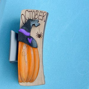 Könnte beinhalten: Haarspange aus Holz mit einem geschnitzten orangefarbenen Kürbis, der einen Hexenhut mit lila Band und eine schwarze Spinne trägt. Das Wort "OCTOBER" ist oben eingraviert. Die Spange ist an einer silbernen Metallbasis befestigt.