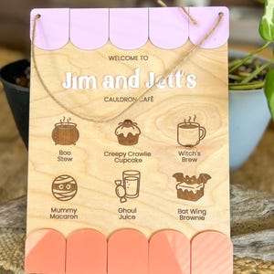 May include: A rectangular wooden sign with a light lavender scalloped top and a coral scalloped bottom. It features the text "WELCOME TO Jim and Jett's CAULDRON CAFÉ" and six engraved food icons with labels: Boo Stew, Creepy Crawlie Cupcake, Witch's Brew, Mummy Macaron, Ghoul Juice, and Bat Wing Brownie. A thin twine string is attached for hanging.