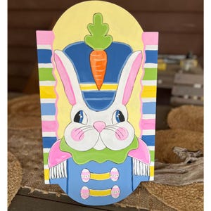 Puede incluir: Decoración de conejo de Pascua pintada a mano. El conejo lleva un sombrero azul con una zanahoria encima y un uniforme azul. El fondo es amarillo con rayas coloridas. El conejo tiene orejas rosas y una nariz rosa.
