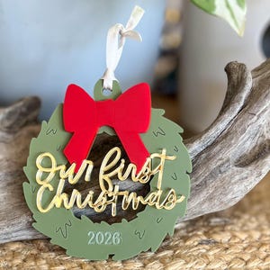May include: A circular olive green Christmas wreath ornament featuring a bright red bow at the top and the gold script text Our First Christmas layered across the center. The year 2026 is engraved at the bottom, and a white ribbon is attached.