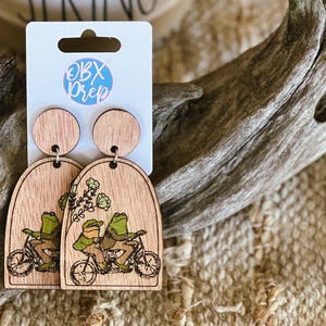 May include: A pair of wooden earrings with a whimsical design of two frogs riding a bicycle. The earrings are shaped like arches and have a wood grain finish. The frogs are green and have black eyes. The bicycle is black and has white wheels. The earrings are labeled "OBX Prep".
