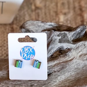May include: A pair of square stud earrings on a white card. The earrings are blue, green, and brown with a stacked design. The card has the text "OBX Prep" in a blue circle.