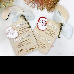 May include: Two wooden Christmas ornaments shaped like keys. Each ornament has a Santa Claus design and the words "Magic Key" and "Santa" printed on it. One ornament has the name "Georgia" printed on it.