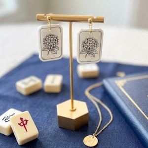 Puede incluir: Un par de pendientes rectangulares con un dibujo lineal en blanco y negro de una flor de hortensia sobre fondo blanco. Los pendientes cuelgan de postes de tono dorado. El diseño imita la forma y el estilo de las fichas de mahjong.