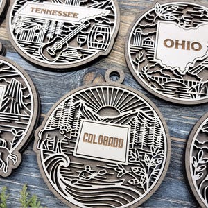 Può includere: Ornamenti in legno circolari con contorni di stati e immagini iconiche. L'ornamento del Tennessee presenta una chitarra e una botte, mentre quello dell'Ohio mostra la forma dello stato. Il Colorado mostra montagne, un fiume e un kayakista.