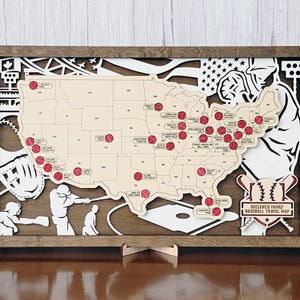 May include: A wooden baseball travel map of the United States, with a detailed cutout design. The map highlights baseball stadiums with red markers. The frame is brown, and the map includes baseball-themed illustrations and text.