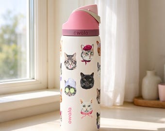 May include: A cream-colored Owala water bottle featuring a pattern of various cat illustrations, including cats wearing berets, spa headbands with cucumber slices, and bows. The bottle has a pink lid with a built-in straw mechanism and carry loop.