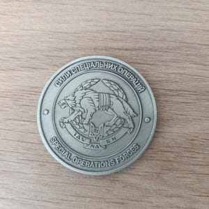 Może przedstawiać: Srebrna moneta z wilkiem w uprzęży pośrodku, otoczona cyrylicą i angielskim tekstem, w tym "SPECIAL OPERATIONS FORCES". Moneta jest okrągła i ma szczegółowy wzór.