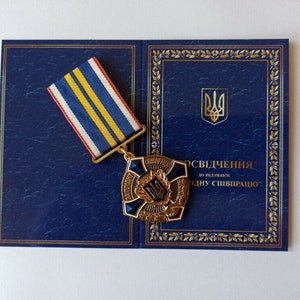 May include: A gold-colored medal with a blue and white ribbon, displayed on a dark blue document with gold trim. The medal features a central emblem and text. The document has a Ukrainian coat of arms.