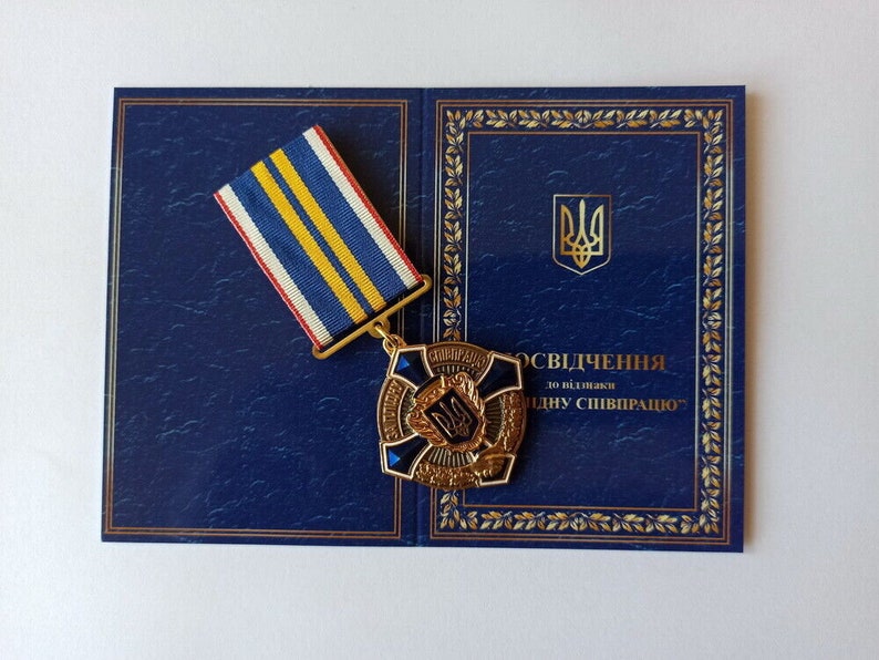 May include: A gold-colored medal with a blue and white ribbon, displayed on a dark blue document with gold trim. The medal features a central emblem and text. The document has a Ukrainian coat of arms.