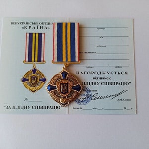 May include: A gold and blue medal with a ribbon, displayed on a certificate. The medal features a cross design with a central emblem and the text "ЗА ПЛІДНУ СПІВПРАЦЮ". The ribbon has blue, yellow, and red stripes. The certificate has Ukrainian text.