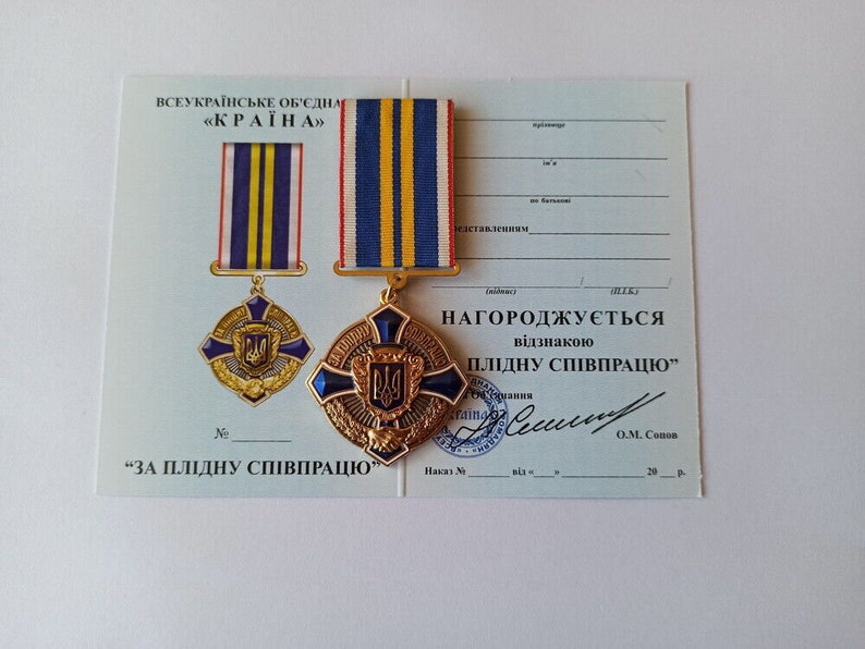 May include: A gold and blue medal with a ribbon, displayed on a certificate. The medal features a cross design with a central emblem and the text "ЗА ПЛІДНУ СПІВПРАЦЮ". The ribbon has blue, yellow, and red stripes. The certificate has Ukrainian text.