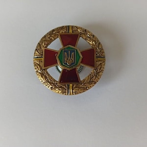 Puede incluir: Medalla circular dorada con diseño de cruz roja. El centro presenta un emblema verde y azul. La medalla está rodeada por una corona de laurel dorada.