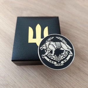 Może przedstawiać: Okrągła, srebrna moneta z czarną obwódką przedstawiająca wilka, gałązki laurowe i tekst. Czarny pojemnik ze złotym emblematem trójzębu znajduje się za monetą. Moneta i pojemnik leżą na jasnej drewnianej powierzchni.