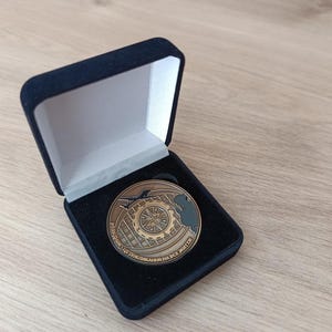 Może przedstawiać: Brązowa moneta pamiątkowa w czarnym aksamitnym pudełku prezentacyjnym. Moneta przedstawia globus z tekstem w języku obcym. Otwarta skrzynka odsłania monetę na białym wnętrzu.