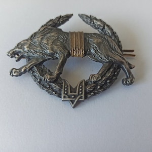 Könnte beinhalten: Ein silberfarbener Metallstift mit einem Wolf, der durch einen Lorbeerkranz springt. Der Wolf hat ein offenes Maul und detailliertes Fell. Der Kranz umgibt ein Dreizack-Symbol. Der Stift hat ein goldfarbenes Band um den Mittelteil des Wolfes.