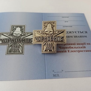 May include: A silver-colored cross-shaped pin with a bell at the top and the word "ЧОРНОБИЛЬ" in the center, along with the dates 1986 and 2021. The pin is on a blue paper with Ukrainian text.