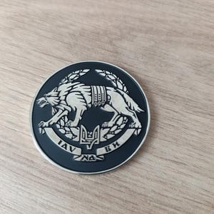 Może przedstawiać: Okrągła, czarno-srebrna moneta z biegnącym wilkiem, otoczonym wieńcem. Moneta ma tekst w języku obcym i symbol trójzębu. Moneta leży na drewnianej powierzchni.