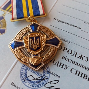 May include: A gold and blue medal with a ribbon, featuring a central emblem and text in Ukrainian. The medal is set against a document with Ukrainian text. The medal has a cross shape with blue accents and a gold emblem.