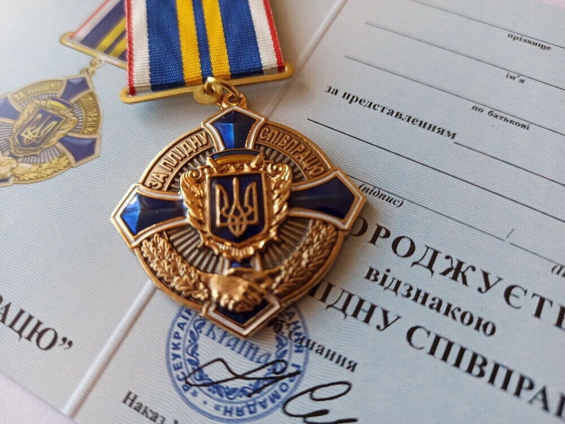 May include: A gold and blue medal with a ribbon, featuring a central emblem and text in Ukrainian. The medal is set against a document with Ukrainian text. The medal has a cross shape with blue accents and a gold emblem.