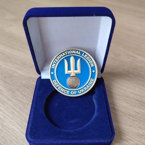 Może przedstawiać: Okrągła, niebiesko-złota moneta Międzynarodowego Legionu Obrony Ukrainy w niebieskim aksamitnym pudełku prezentacyjnym. Moneta przedstawia symbol trójzębu oraz napisy "INTERNATIONAL LEGION" i "DEFENCE OF UKRAINE".