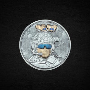 Puede incluir: Una moneda de plata con un relieve detallado de un soldado con equipo de combate. El soldado lleva un casco, gafas azules y auriculares. Dos objetos con forma de oso estilizados de color beige están encima del casco. La moneda está sobre un fondo negro.