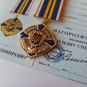 May include: A gold-toned medal with a blue and gold ribbon, featuring a central emblem and the words "ЗА ПЛІДНУ СПІВПРАЦЮ". The medal is set against a light blue document with Ukrainian text and a seal.