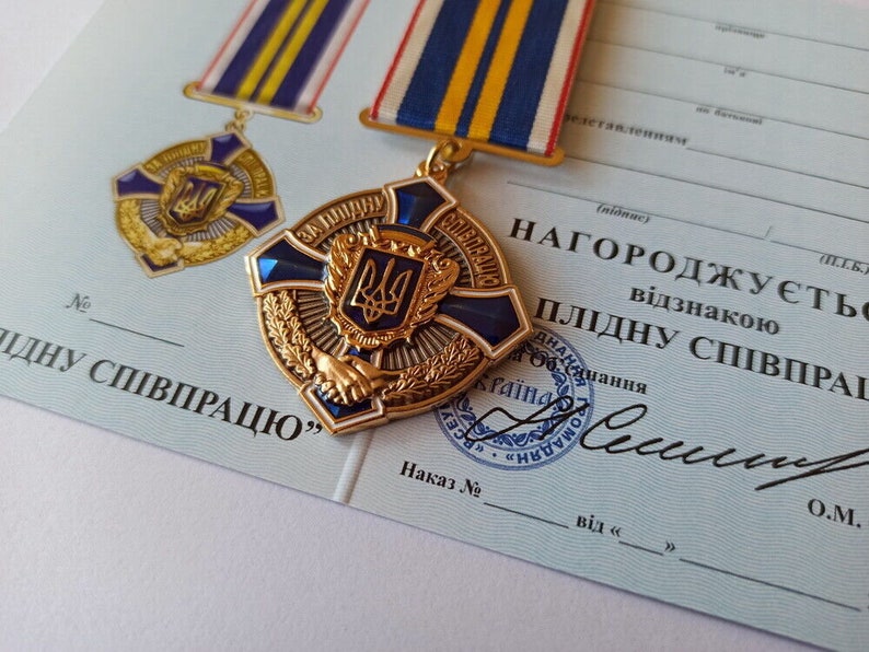 May include: A gold-toned medal with a blue and gold ribbon, featuring a central emblem and the words "ЗА ПЛІДНУ СПІВПРАЦЮ". The medal is set against a light blue document with Ukrainian text and a seal.