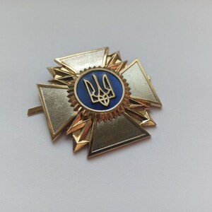 Könnte beinhalten: Eine goldfarbene, kreuzförmige Medaille mit einem blauen, kreisförmigen Zentrum, das ein goldenes Dreizack-Emblem zeigt. Die Medaille hat ein Sternmuster um das Zentrum und ist vor einem weißen Hintergrund platziert.