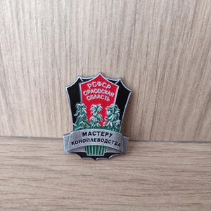 Może przedstawiać: Vintage metalowa przypinka w kształcie tarczy. Przypinka zawiera tekst "РСФСР ОРАОВСКАЯ ОБЛАСТЬ" w kolorze czerwono-białym oraz "МАСТЕРУ КОНОПЛЕВОДСТВА" w kolorze srebrnym. Projekt zawiera zielono-białe ilustracje roślin.