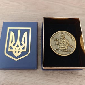 Może przedstawiać: Niebieskie pudełko ze złotym herbem w kształcie trójzębu obok otwartego pudełka ze złotą monetą. Moneta przedstawia żołnierza z karabinem i ukraińskim tekstem. Moneta znajduje się we wnętrzu z czarnego aksamitu.