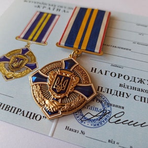 May include: A gold-colored medal with blue enamel and a central emblem, accompanied by a matching ribbon. The medal is on a light blue document with Ukrainian text and a seal. The medal features a cross design and a wreath.