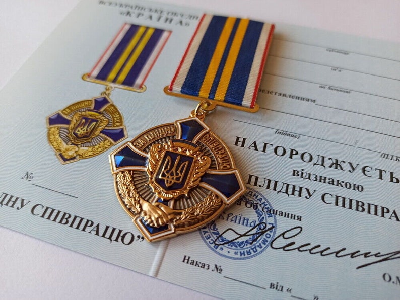 May include: A gold-colored medal with blue enamel and a central emblem, accompanied by a matching ribbon. The medal is on a light blue document with Ukrainian text and a seal. The medal features a cross design and a wreath.