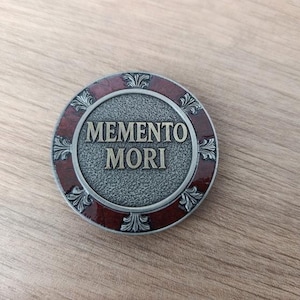 Może przedstawiać: Okrągła, metalowa moneta w stylu antycznym z napisem "MEMENTO MORI". Moneta ma teksturowane, szare centrum, ciemnoczerwony obramowanie i dekoracyjne zdobienia. Leży na drewnianej powierzchni.
