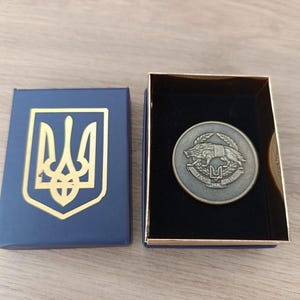 Może przedstawiać: Niebieskie pudełko ze złotym herbem trójzębu i srebrną monetą z motywem wilka i trójzębu. Moneta jest prezentowana w czarnym pudełku wyłożonym aksamitem ze złotym obramowaniem.