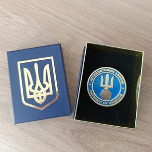 Może przedstawiać: Niebieskie pudełko ze złotym emblematem i przypinką z napisem "INTERNATIONAL LEGION DEFENCE OF UKRAINE" oraz symbolem trójzębu. Przypinka jest niebiesko-złota, a pudełko otwarte na drewnianej powierzchni.