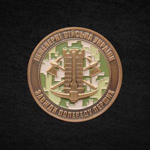 May include: A bronze-colored coin with a central design featuring a castle, crossed axes, and a pickaxe. The coin has text in a circular pattern around the edge, with a green and white camouflage background.