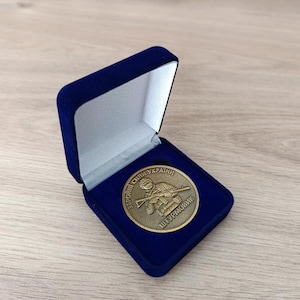 Może przedstawiać: Brązowy medal w niebieskim aksamitnym pudełku prezentacyjnym. Medal przedstawia żołnierza z karabinem i tekstem w obcym języku. Pudełko jest otwarte, ukazując medal.