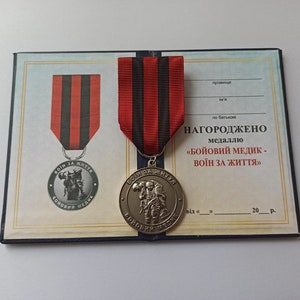 Peut inclure: Une médaille dorée avec un ruban rouge et noir, présentée sur un certificat. La médaille présente une image en relief et le texte "BOЙOBИЙ MEДИK - BOIН 3A ЖИTTЯ". Le certificat contient du texte en ukrainien.