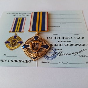 May include: A gold-colored medal with a blue and gold ribbon, featuring a crest with a trident. The medal is displayed on a certificate with Ukrainian text, including the phrase "For fruitful cooperation."