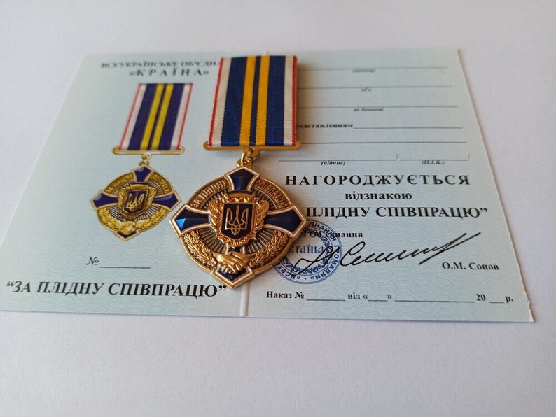 May include: A gold-colored medal with a blue and gold ribbon, featuring a crest with a trident. The medal is displayed on a certificate with Ukrainian text, including the phrase "For fruitful cooperation."
