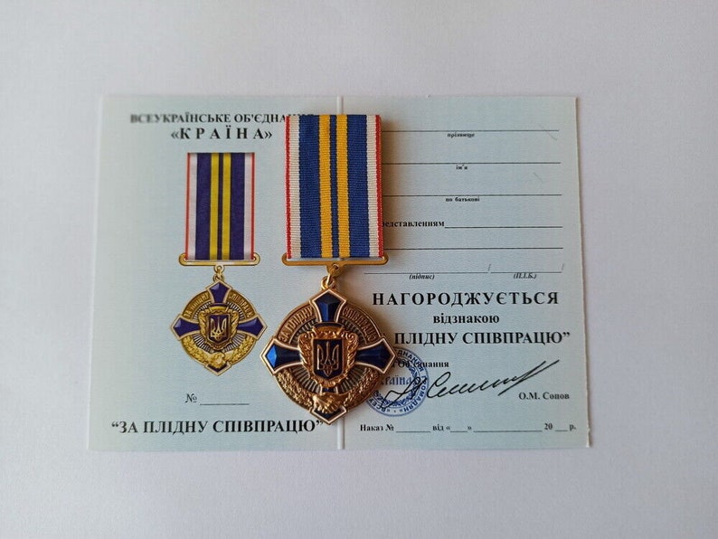 May include: A Ukrainian medal with a blue and yellow striped ribbon, a gold-colored cross-shaped emblem, and the text "ЗА ПЛІДНУ СПІВПРАЦЮ" on a certificate. The medal features a blue and gold emblem.