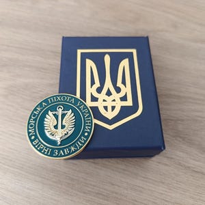 Puede incluir: Un emblema militar redondo, verde azulado y dorado, con texto ucraniano y un diseño central de ancla y alas. Junto a él, una caja azul marino con un escudo de armas ucraniano dorado.