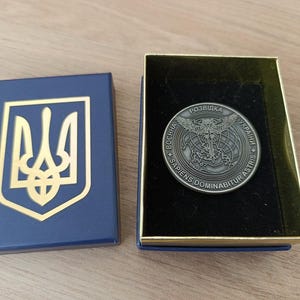 Może przedstawiać: Niebieskie pudełko ze złotym emblematem i moneta w pudełku ze złotymi obramowaniami. Moneta ma szczegółowy wzór z tekstem w obcym języku. Pudełka znajdują się na jasnobrązowej powierzchni.
