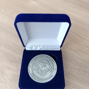 Może przedstawiać: Srebrna moneta z napisem "SPECIAL OPERATIONS FORCES" i tekstem cyrylicą, prezentowana w niebieskim aksamitnym pudełku. Moneta przedstawia szczegółowy emblemat stworzenia podobnego do wilka. Pudełko jest otwarte, ukazując monetę.